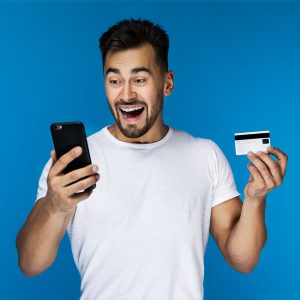surprised smiling attractive guy is watching on the cell phone screen and holding credit card in the hand